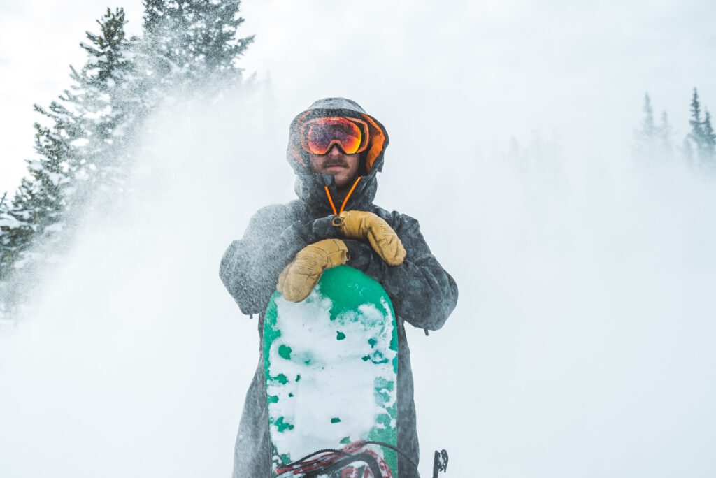 Man standing in snow, wearing winter gear, holding a snowboard, and equipped with ski mittens.