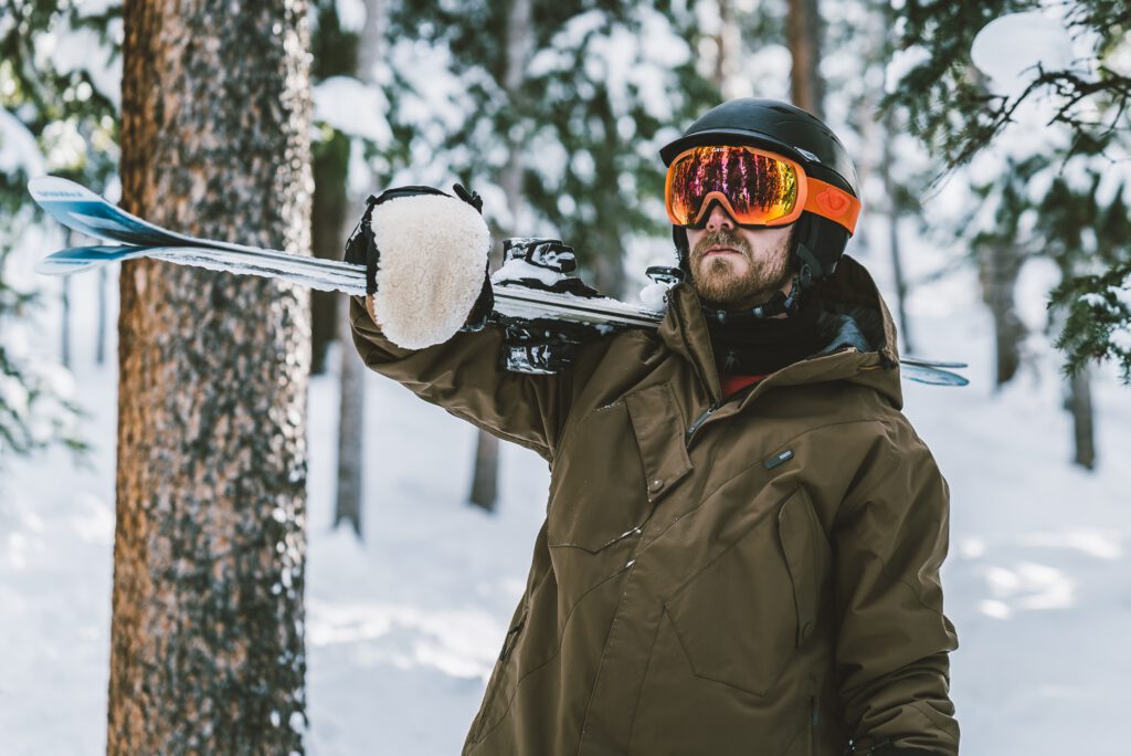 A man standing outdoors in winter clothing, holding skis and wearing ski mittens.