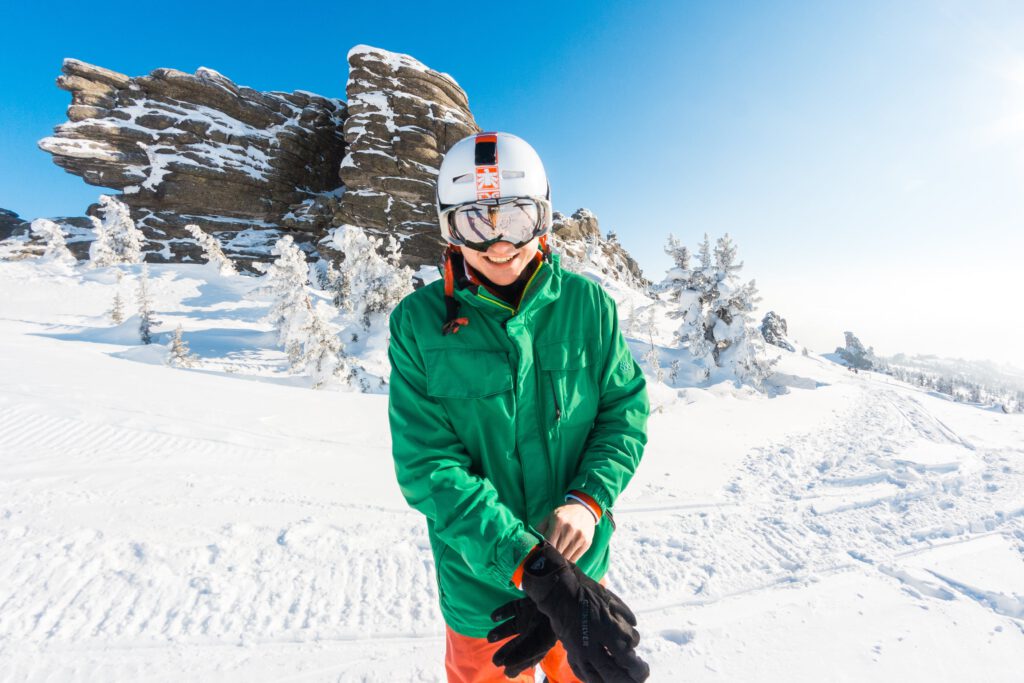 Man in green jacket and ski glasses putting on ski gloves, preparing for winter sports.